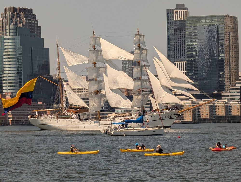 A large white sailing ship with multiple sails and a colorful flag is on the water near a city, while several people paddle yellow and red kayaks in the foreground.
