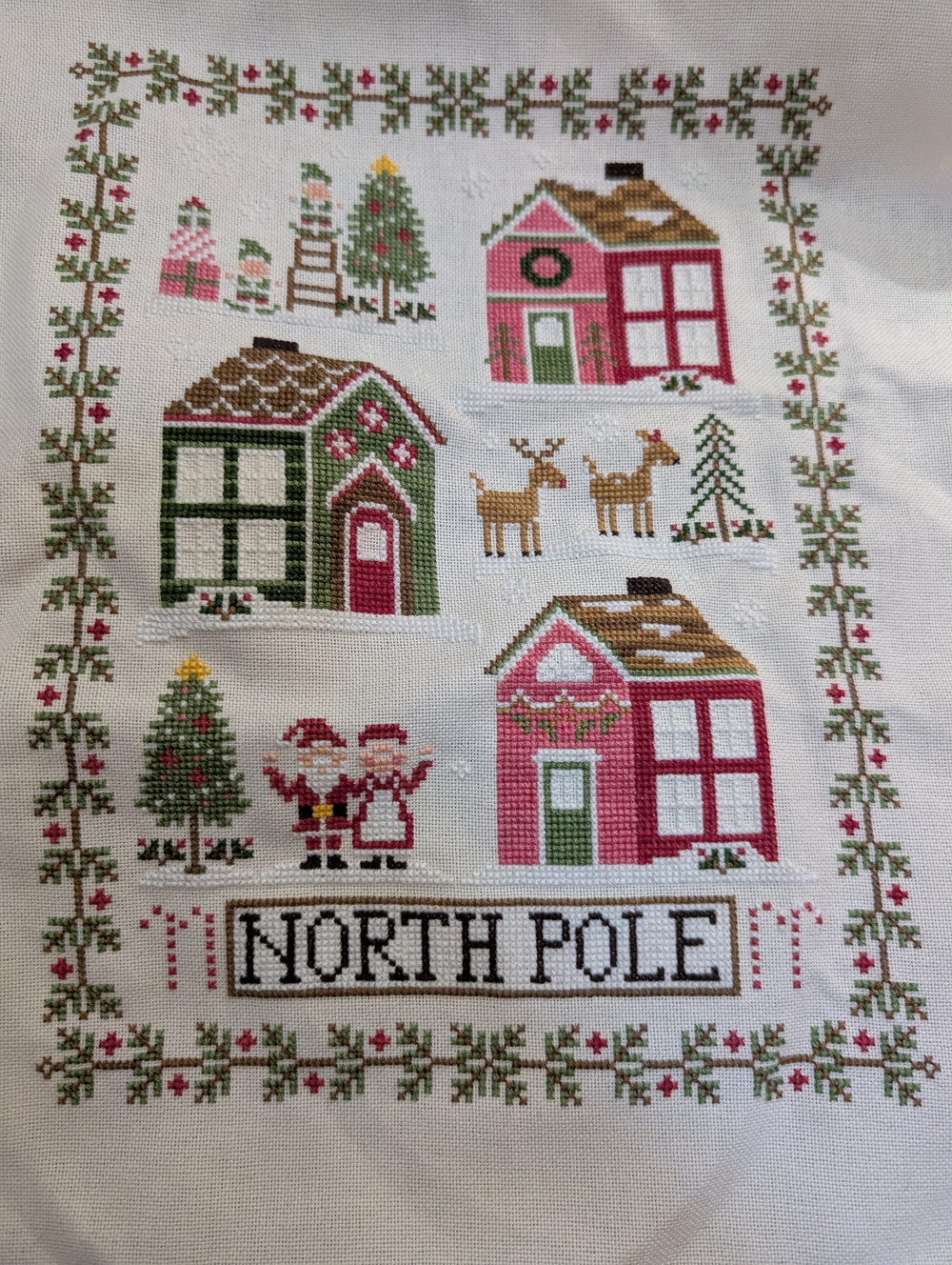 A cross-stitch artwork depicts festive houses, reindeer, Santa and Mrs. Claus, and the words NORTH POLE surrounded by a decorative border.