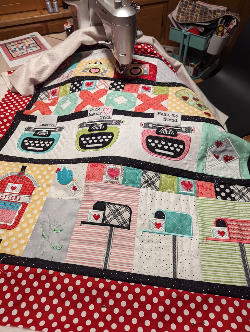 A colorful patchwork quilt featuring retro-style typewriters and mailboxes is being worked on with a sewing machine.