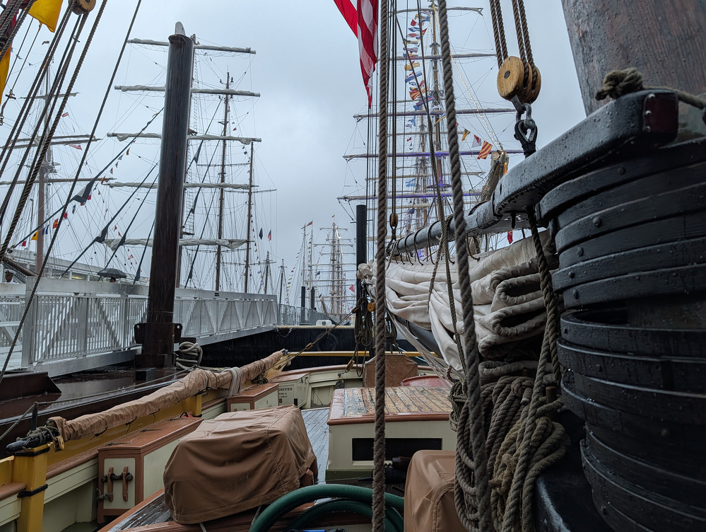 A docked sailing ship with tightly furled sails and numerous ropes is surrounded by other tall ships with multiple masts against a cloudy sky.