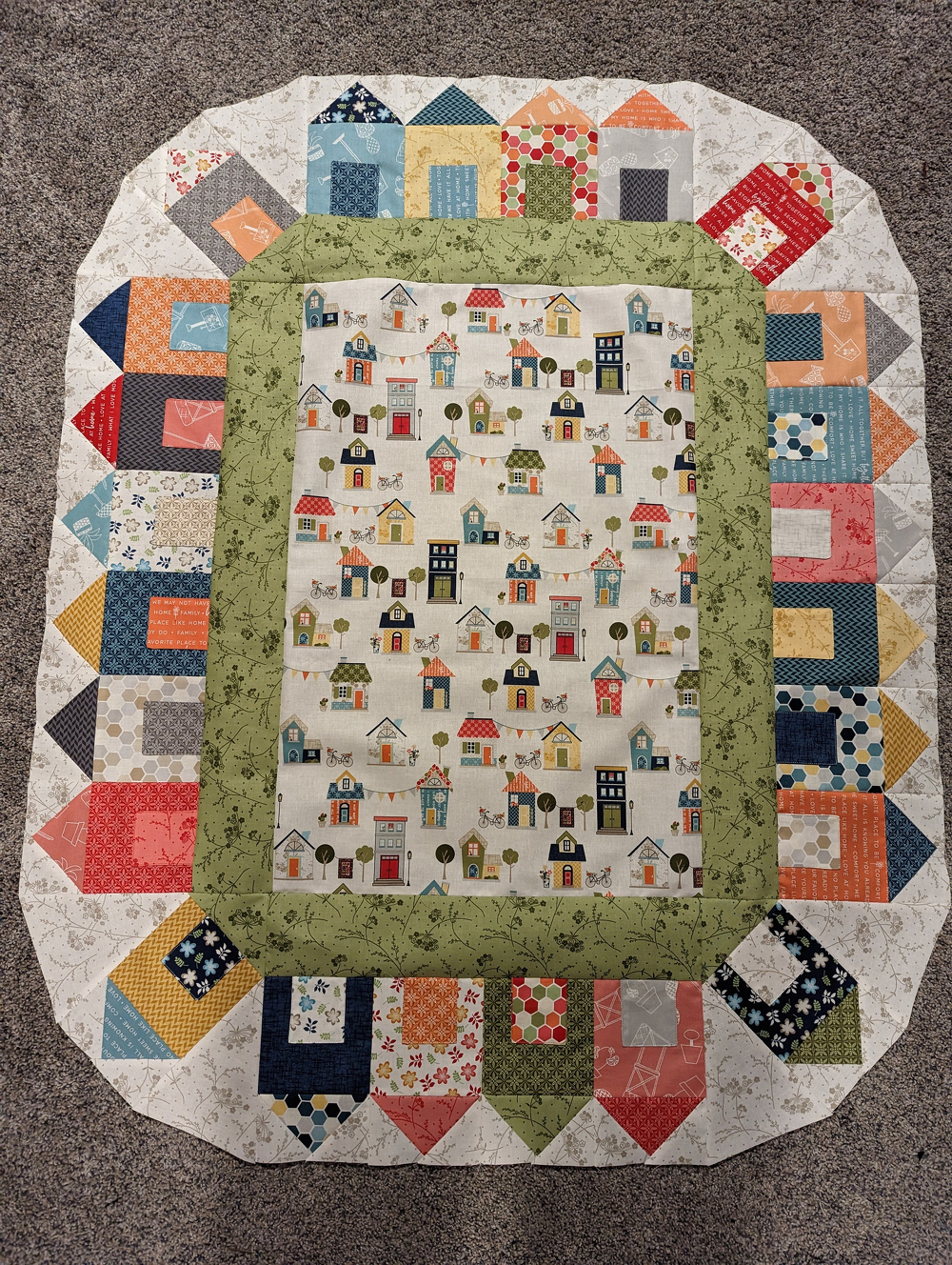 A patchwork quilt features various colorful patterns with a central design of small houses surrounded by a green border and intricate geometric motifs.