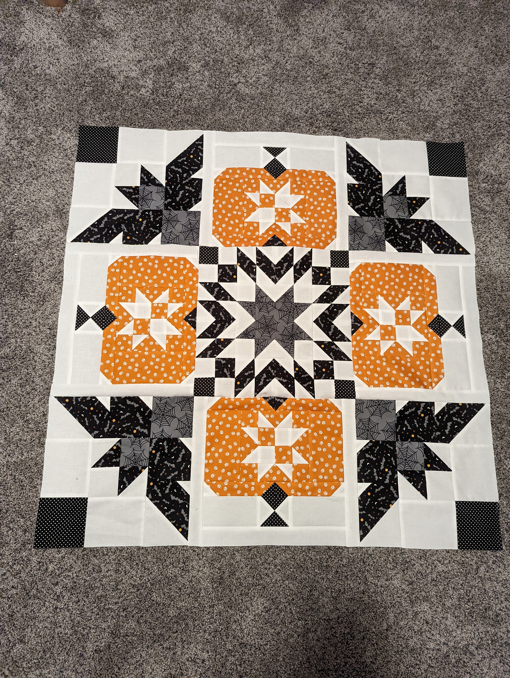A quilt block featuring geometric patterns with orange, black, and white fabric designs, resembling pumpkins and stars, is laid out on a carpeted floor.