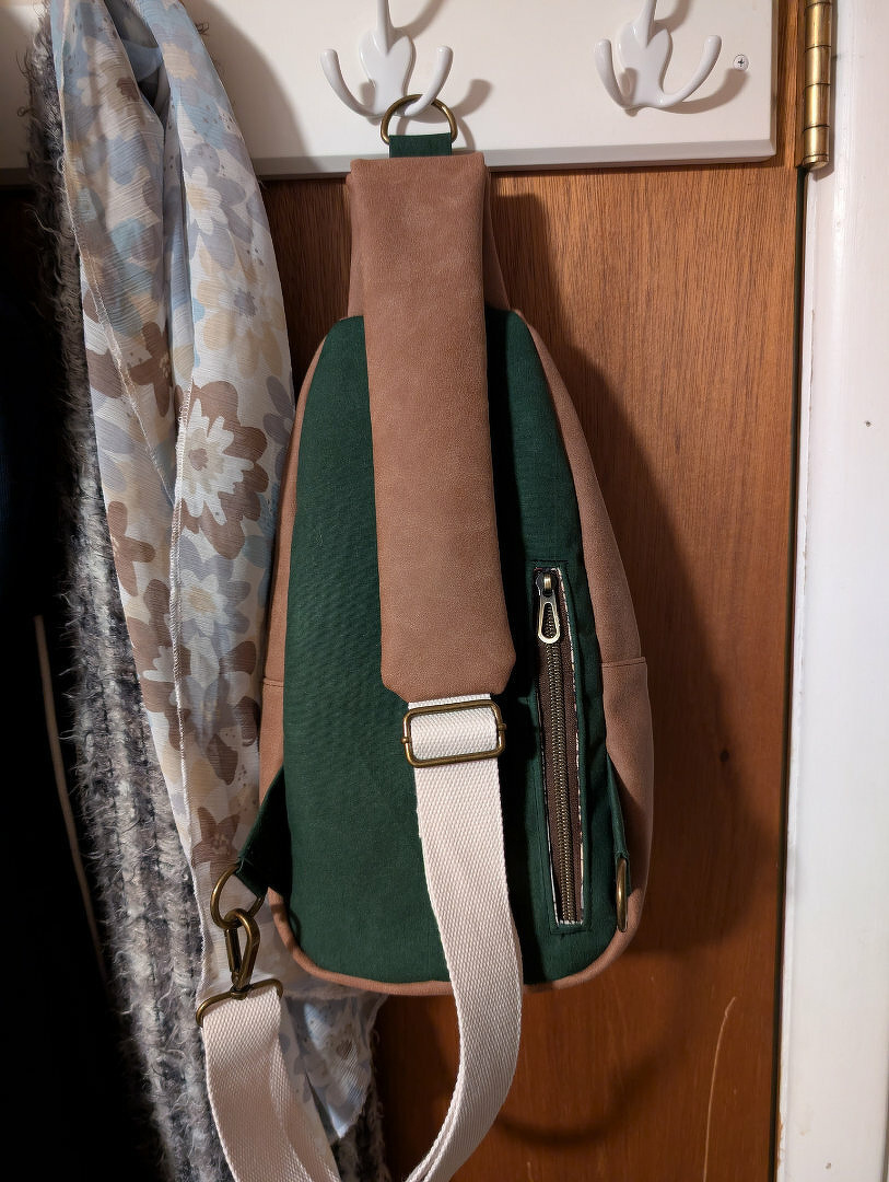 A backpack with a green body and brown accents is hanging from hooks on a wooden door, next to a floral-patterned scarf.