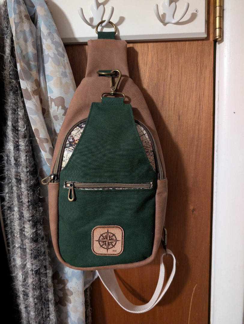A stylish sling bag with green and brown colors hangs on a wooden door, featuring a front zipper pocket and a compass design patch.