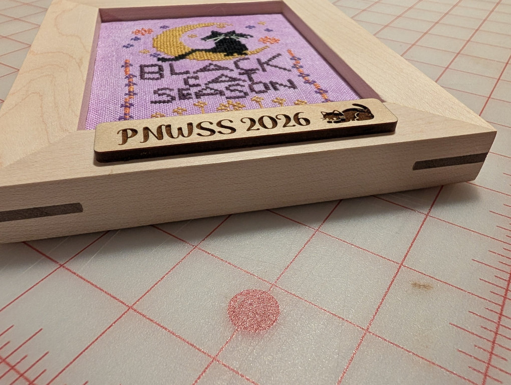 A framed cross-stitch artwork with a witch on a crescent moon and the words Black Season is displayed on a cutting mat, with a wooden plaque labeled PNWSS 2026 featuring a cat illustration.