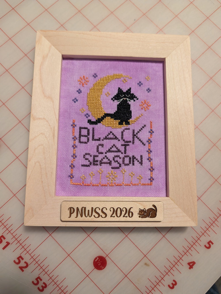 A cross-stitched design features a black cat sitting on a crescent moon with the words BLACK CAT SEASON, framed in a wooden frame labeled PNWSS 2026 with a small cat illustration.