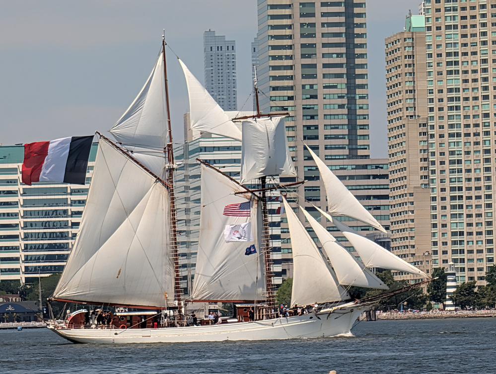 A tall ship with multiple white sails is sailing on the water near a cityscape with high-rise buildings in the background.