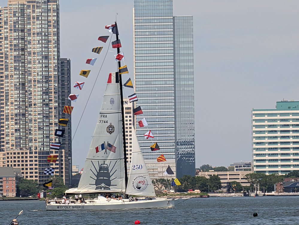 A sailboat decorated with multiple colorful signal flags is sailing near a city waterfront with tall buildings in the background.