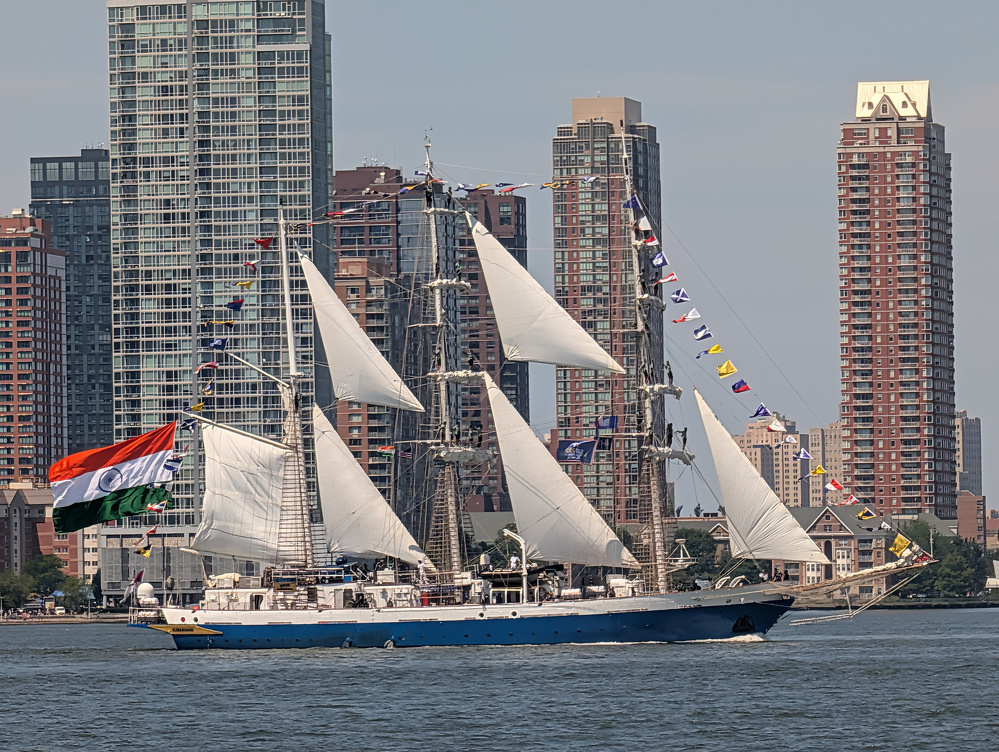 A large sailing ship with white sails and colorful flags is sailing on the water in front of a cityscape with tall buildings.