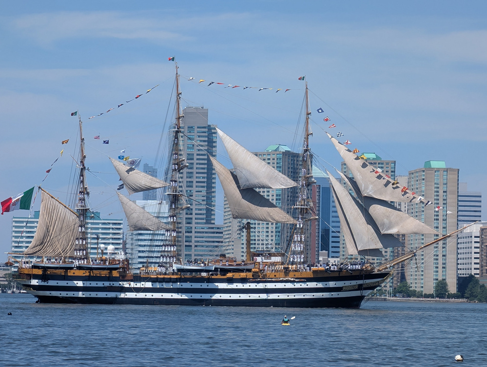 A large, vintage sailing ship with multiple masts and colorful flags is floating on the water in front of a city skyline.