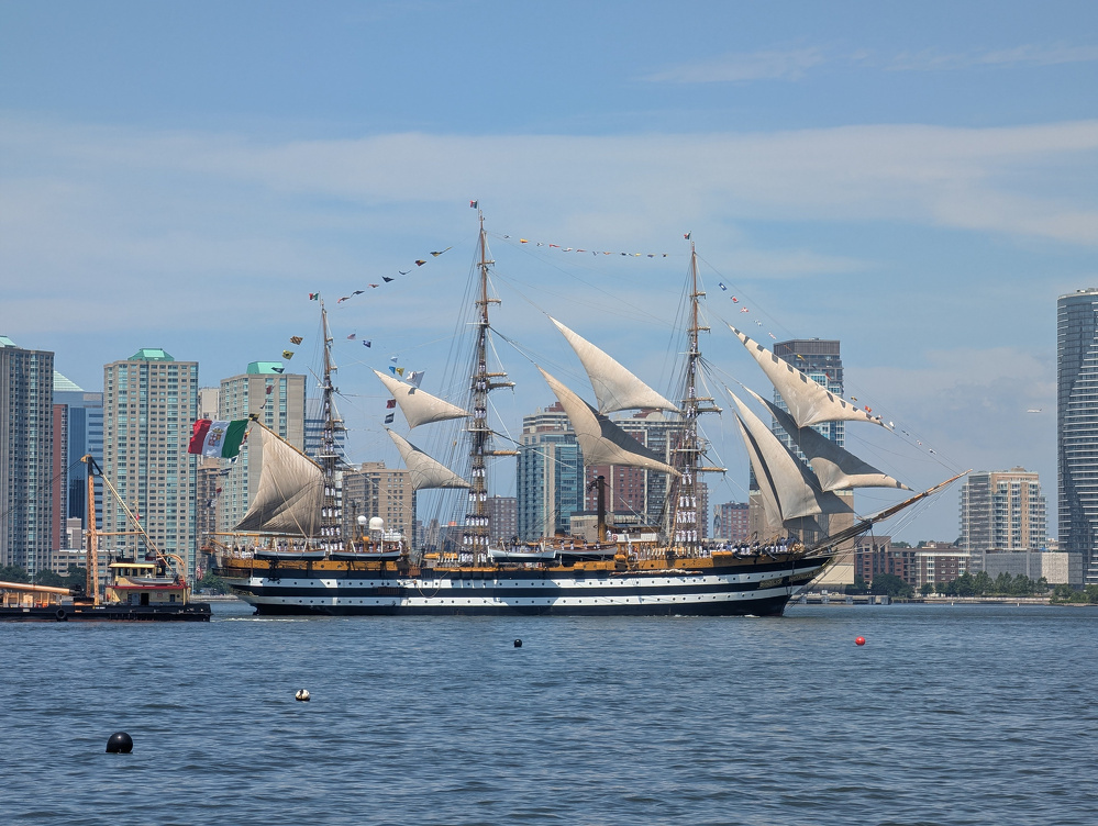 A large, multi-masted sailing ship with white sails is floating on calm water near a city skyline with modern high-rise buildings.