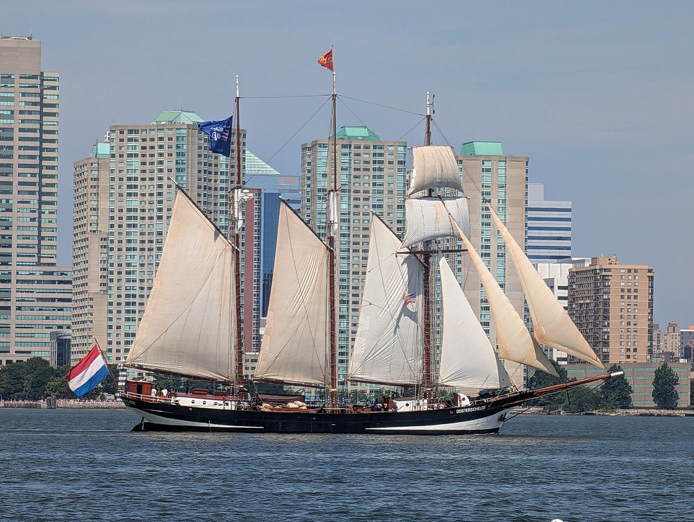 A large sailing ship with multiple white sails is moving on the water against a backdrop of tall city buildings.
