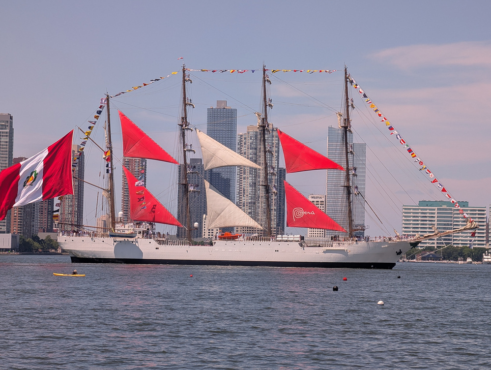A large white sailing ship with red and white sails and a Peruvian flag is floating on a body of water with a city skyline in the background.