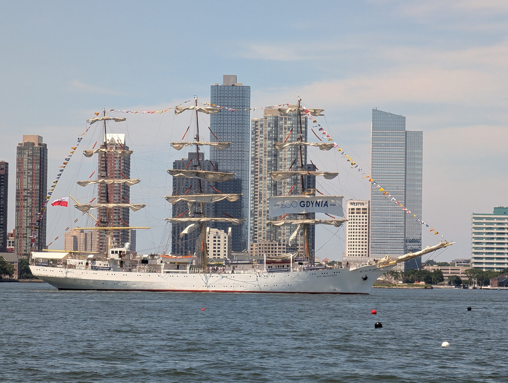A large, white sailing ship with multiple masts and colorful flags is docked near a cityscape with tall buildings.