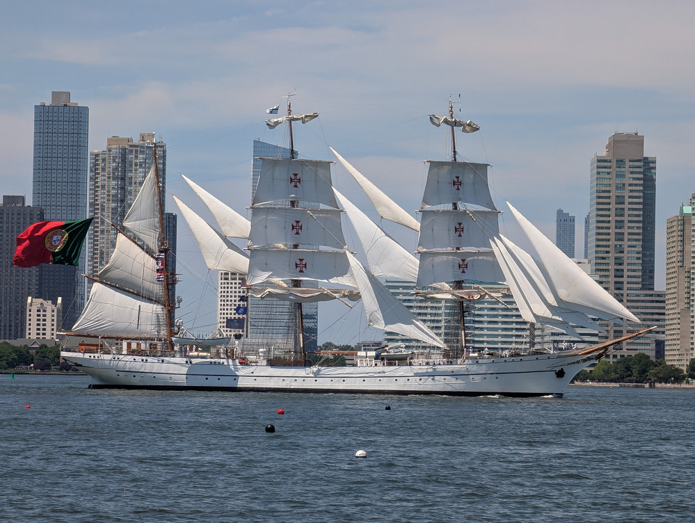 A large white sailing ship with multiple sails raised is sailing on a body of water with a city skyline in the background.