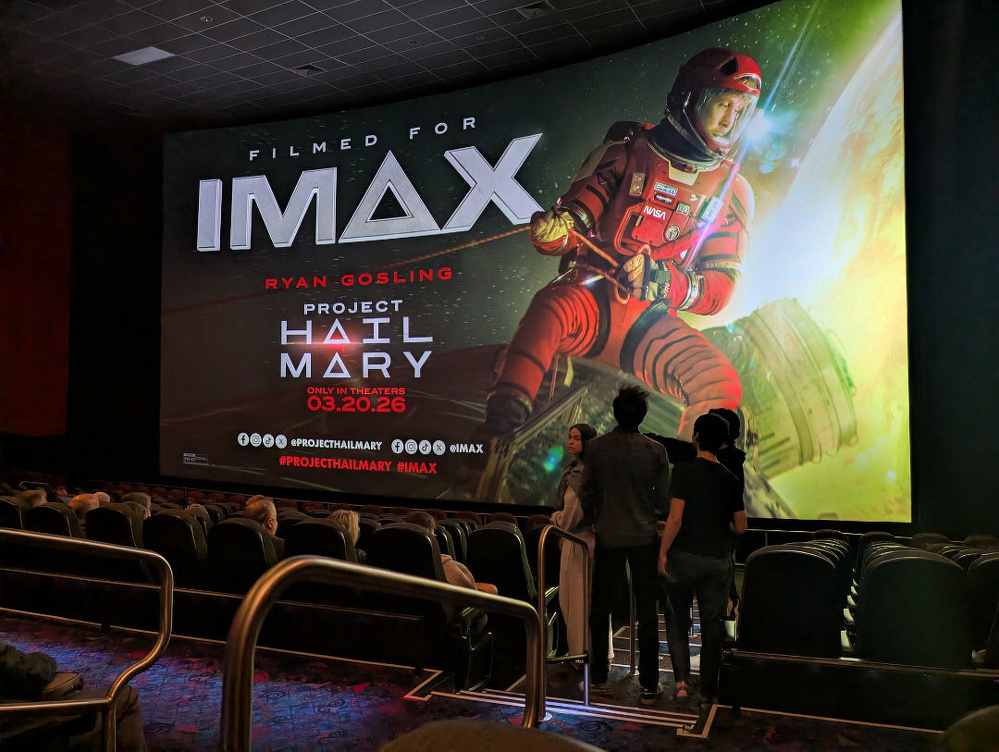 A movie theater screen displays a poster for Project Hail Mary, featuring an astronaut and promoting its IMAX release, with people seated and standing in front of it.