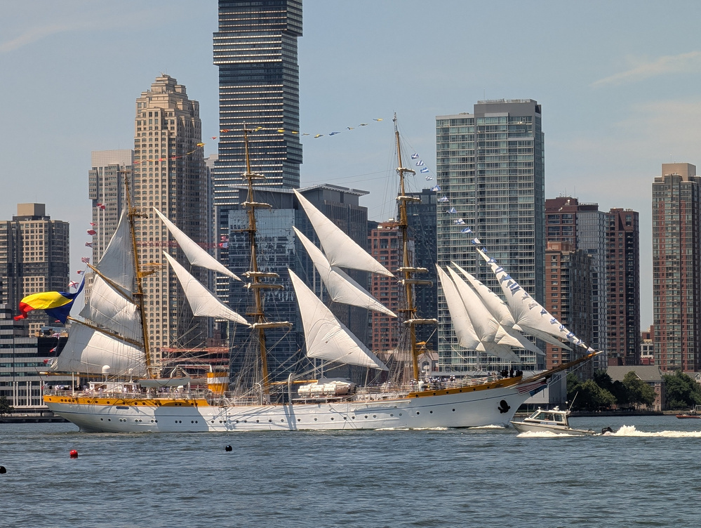 A large white sailing ship with multiple sails raised is gliding on the water in front of a city skyline with tall buildings.