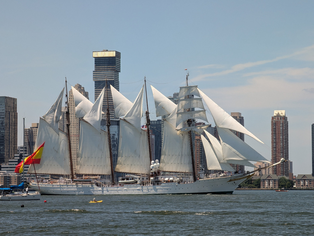 A large, multi-masted sailing ship with white sails is navigating a body of water near a cityscape with tall buildings.