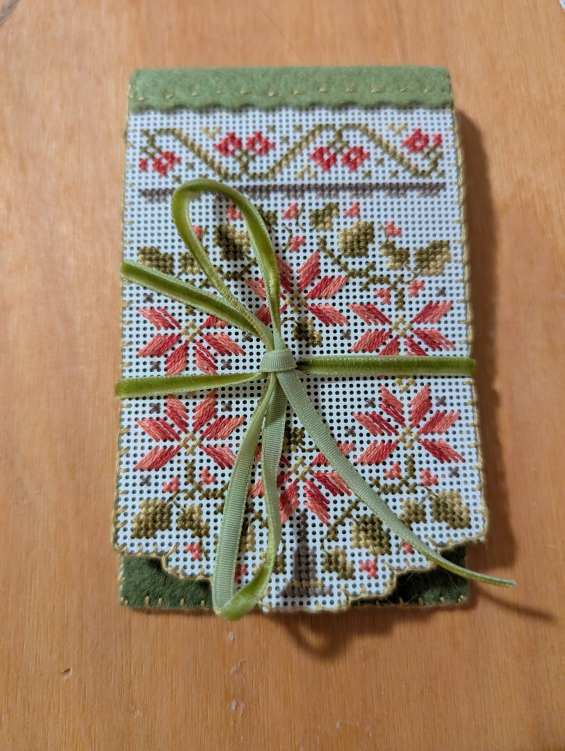 A rectangular, needlepoint fabric with red and green floral patterns is tied with a green ribbon on a wood surface.