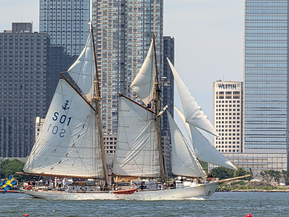 A large white sailboat with multiple sails is sailing on the water with a city skyline of tall buildings in the background.