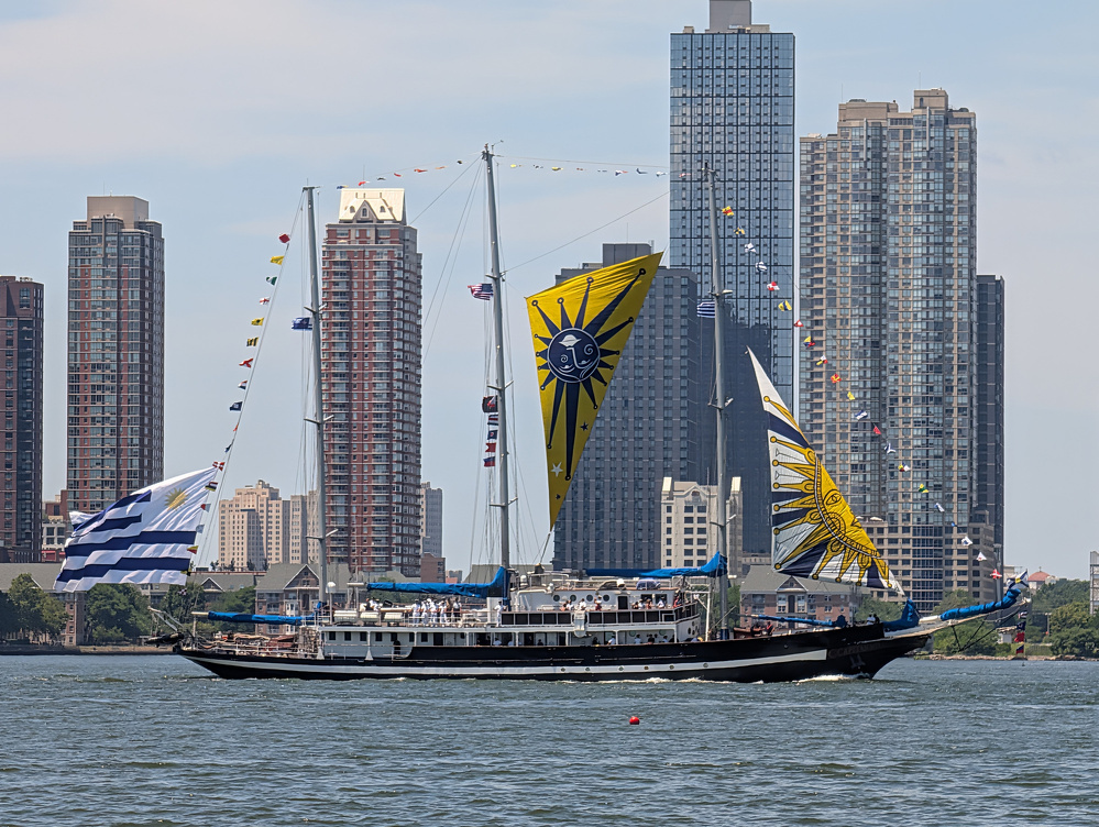 A large sailboat with colorful sun-themed sails and flags is sailing on the water with tall city buildings in the background.