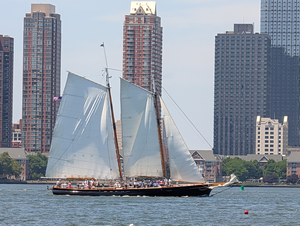 A large sailboat with white sails is sailing on a body of water near a cityscape with tall buildings in the background.