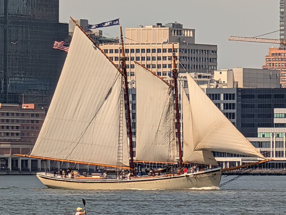 A large sailboat with multiple sails is sailing on a body of water near a city with tall buildings in the background.