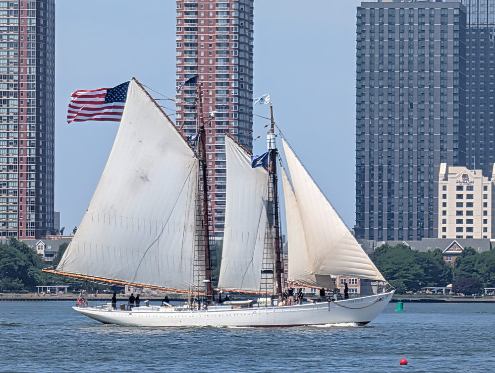 A white sailing ship with its sails fully unfurled and an American flag flying at the stern is sailing on a body of water with tall buildings in the background.