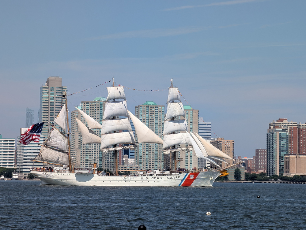A U.S. Coast Guard tall ship with white sails is sailing on the water near a city skyline with multiple high-rise buildings.