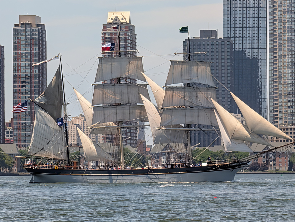 A large, historic sailing ship with multiple white sails is navigating a body of water in front of a city skyline with tall buildings.