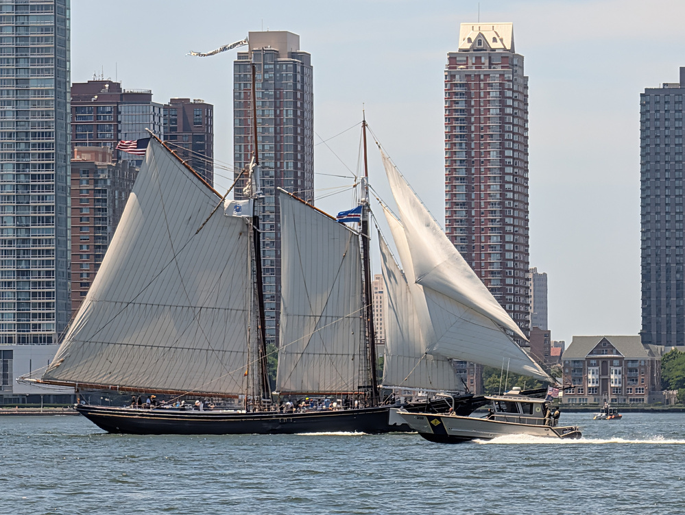 Two sailboats with their sails unfurled are sailing on a body of water against a backdrop of tall urban buildings.