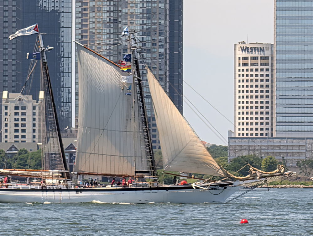 A sailboat with its sails unfurled is navigating a body of water near a cityscape with tall buildings.
