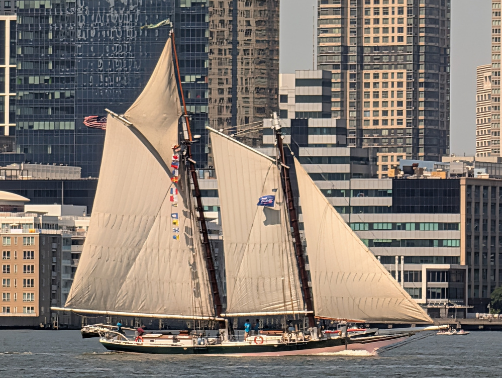 A sailboat with large beige sails is sailing on the water in front of a densely packed urban skyline with various modern buildings.