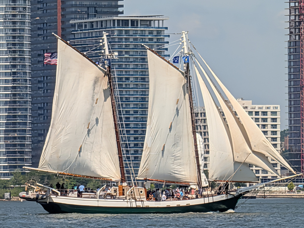 A classic sailing ship with white sails is moving on the water against a backdrop of tall modern buildings.