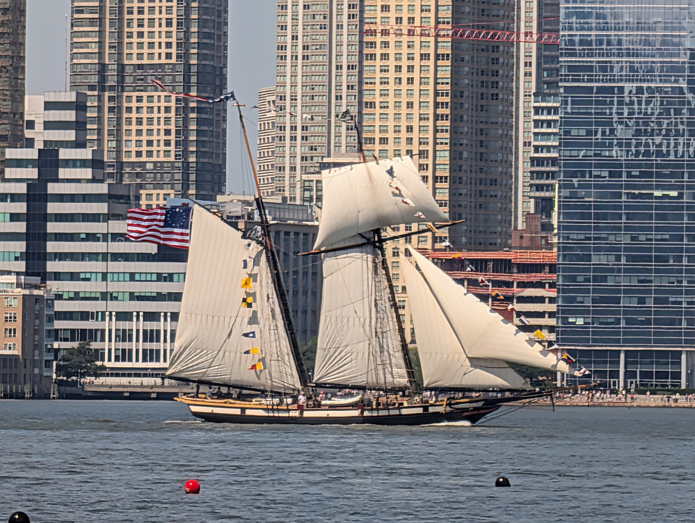 A large sailing ship with white sails and an American flag is cruising on the water with a backdrop of tall city skyscrapers.