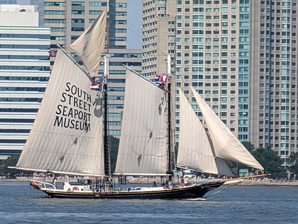 A sailboat with large white sails bearing the text South Street Seaport Museum is sailing on the water near a cityscape with tall buildings.