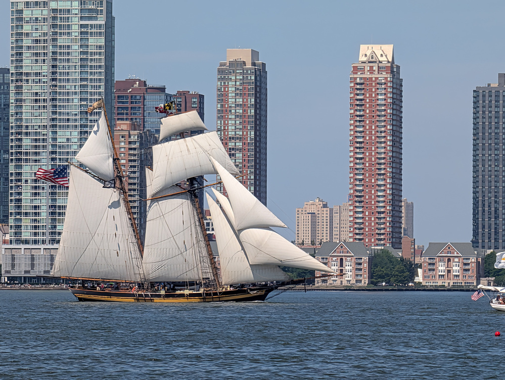 A large sailing ship with white sails is gliding on the water near a cityscape of tall buildings.