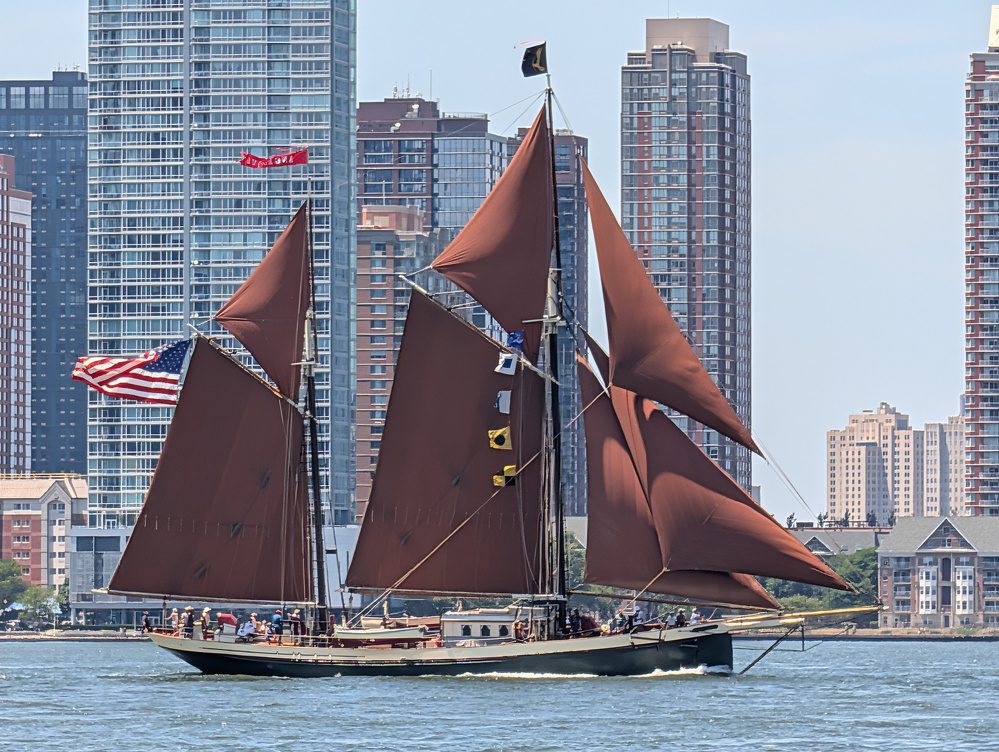 A traditional sailing ship with brown sails and an American flag is sailing on the water near a city skyline with tall buildings.