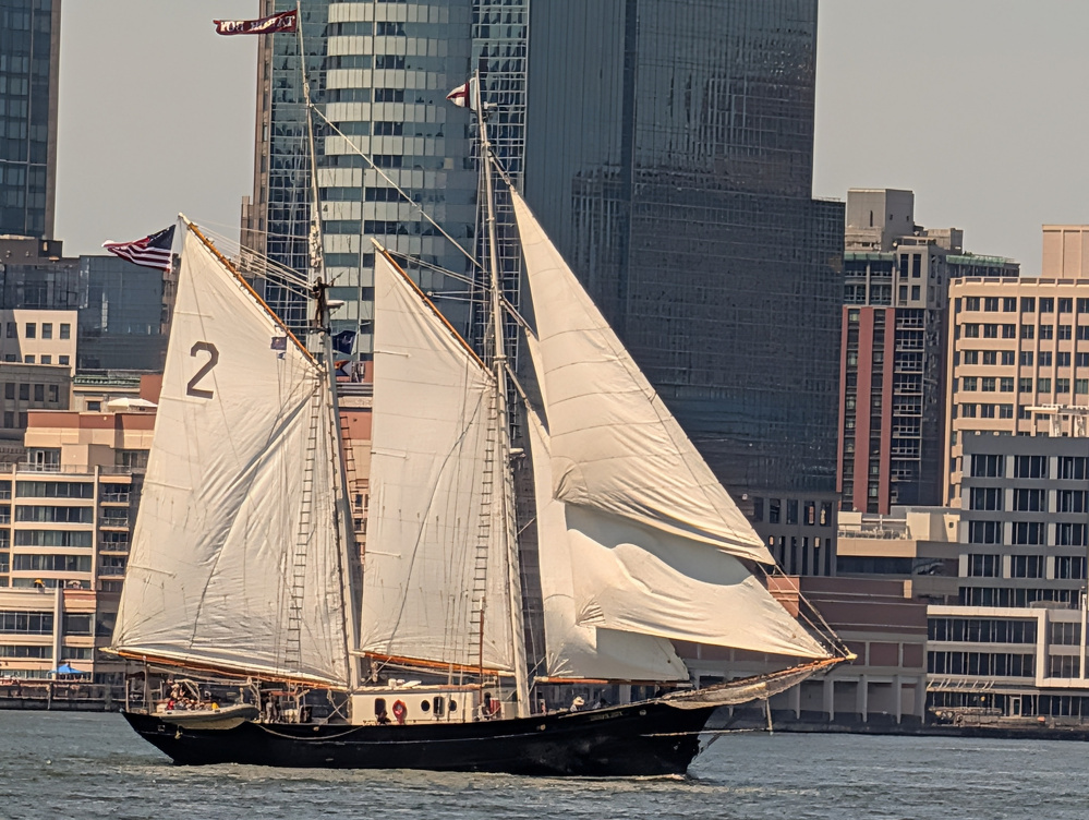 A large sailboat with multiple white sails glides on the water against a backdrop of urban buildings and skyscrapers.