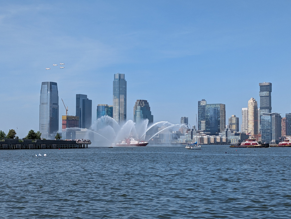 A city skyline with water in the foreground features a fireboat spraying water fountains and four planes flying in formation above.