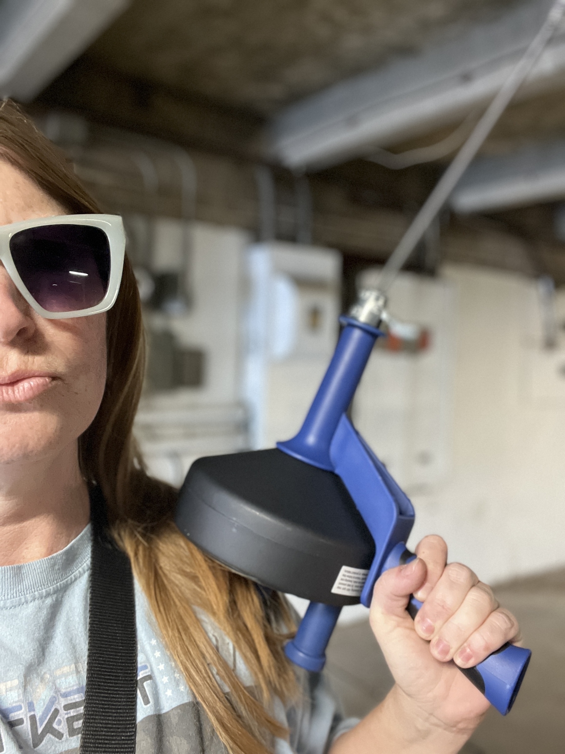 A woman wearing white sunglasses holds a blue and black plumbing snake in her left hand. She is wearing a light blue shirt with a black strap around her neck. The background is out of focus.