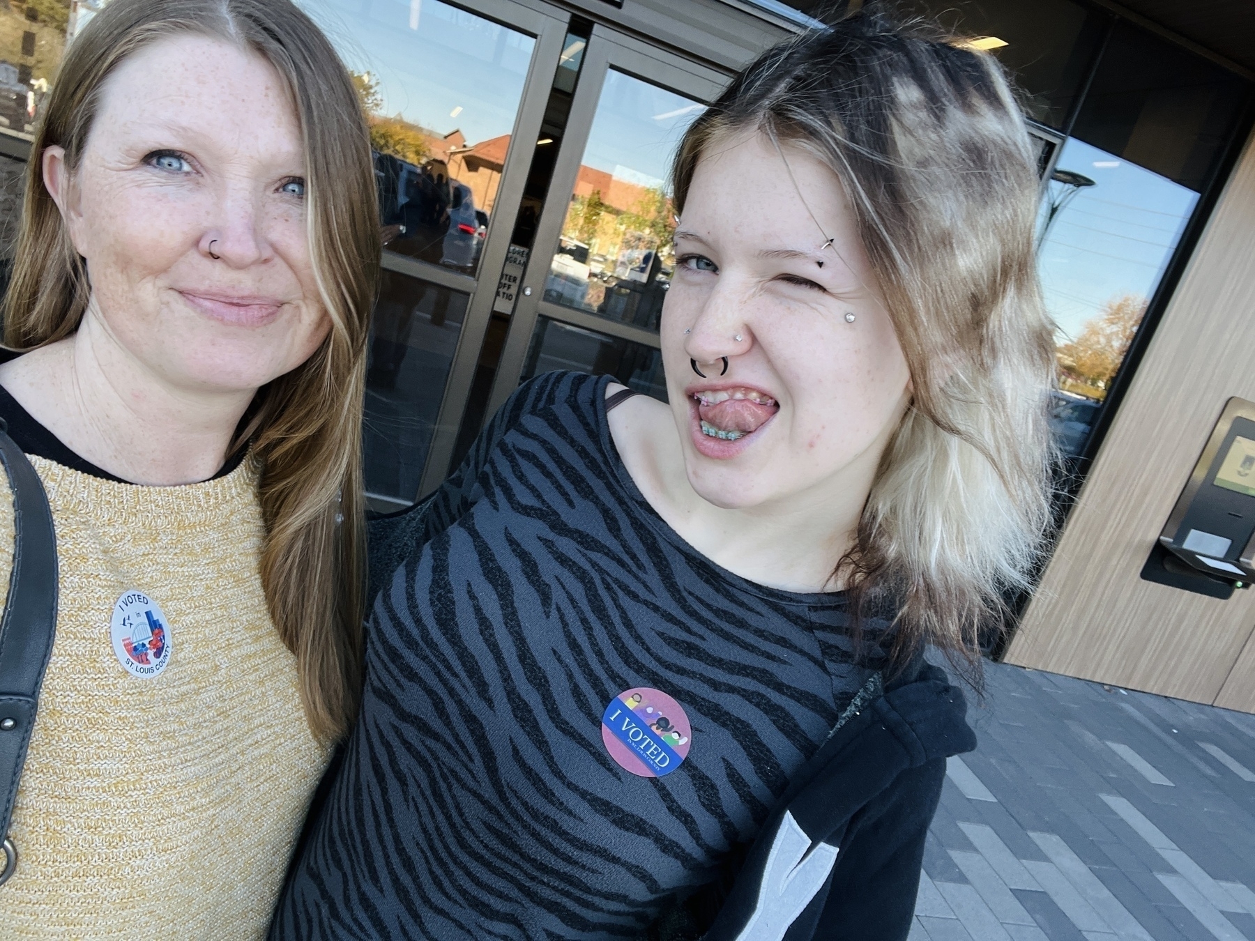 Two women - a mother and daughter - wearing voting stickers and standing outside a polling place. The mother is smiling, the daughter is winking.