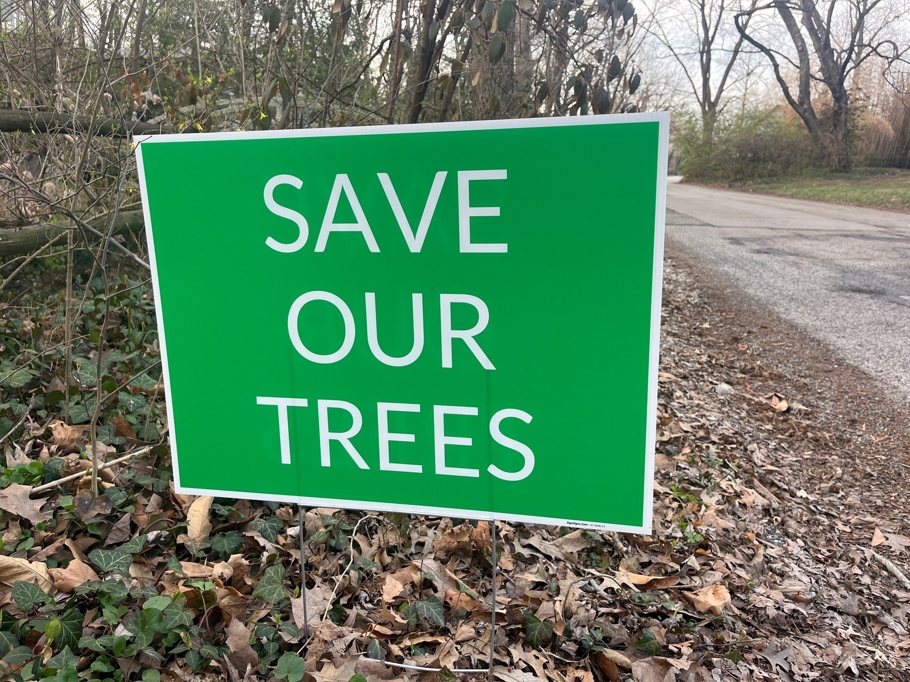 A green sign that says Save Our Trees on the side of a narrow road