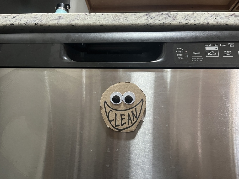 On the front of a stainless steel dishwasher, there’s a small cardboard circle with googly eyes and the word CLEAN inside a grinning mouth, drawn with black marker