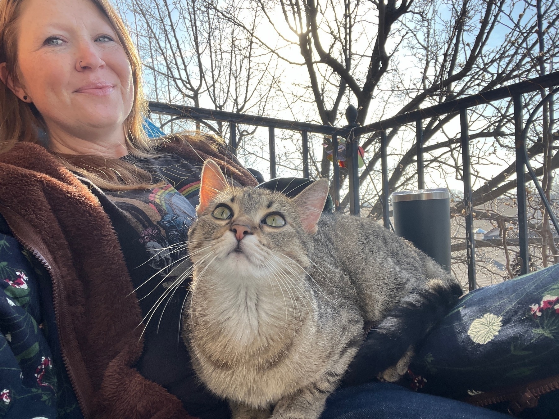 A woman sits on a balcony holding a small gray tabby cat with wide eyes 