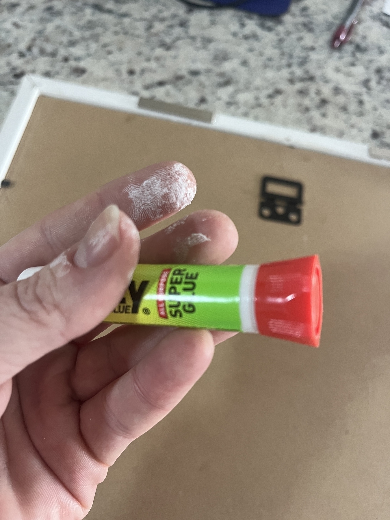 A hand is holding a bottle of superglue with visible dried superglue on three fingers. The background is a counter with the back of a picture.