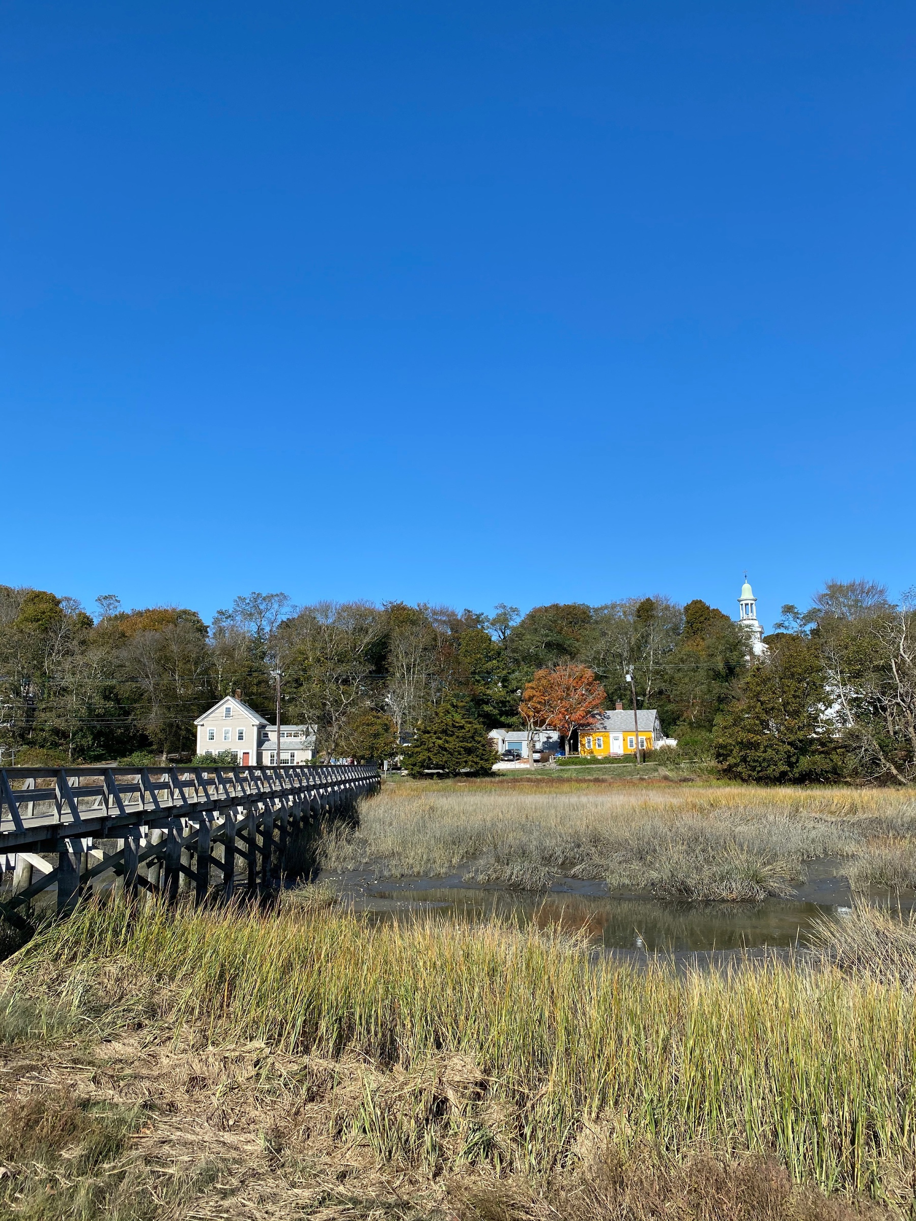 View of a wooden bridge over a creek, with buildings and a white church steeple on the far bank.