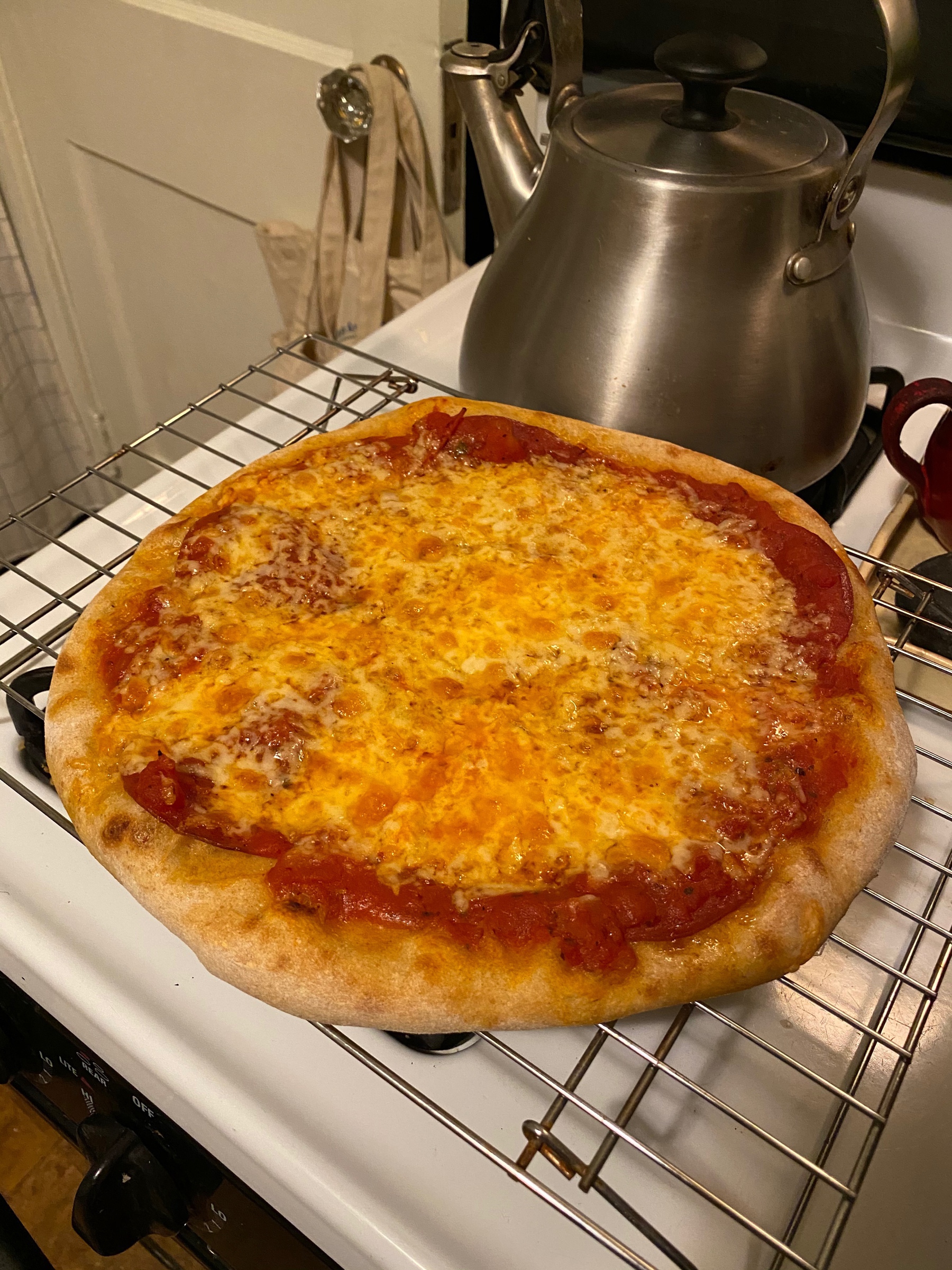 A small pizza on a cooling rack on a stove with a kettle in the background.
