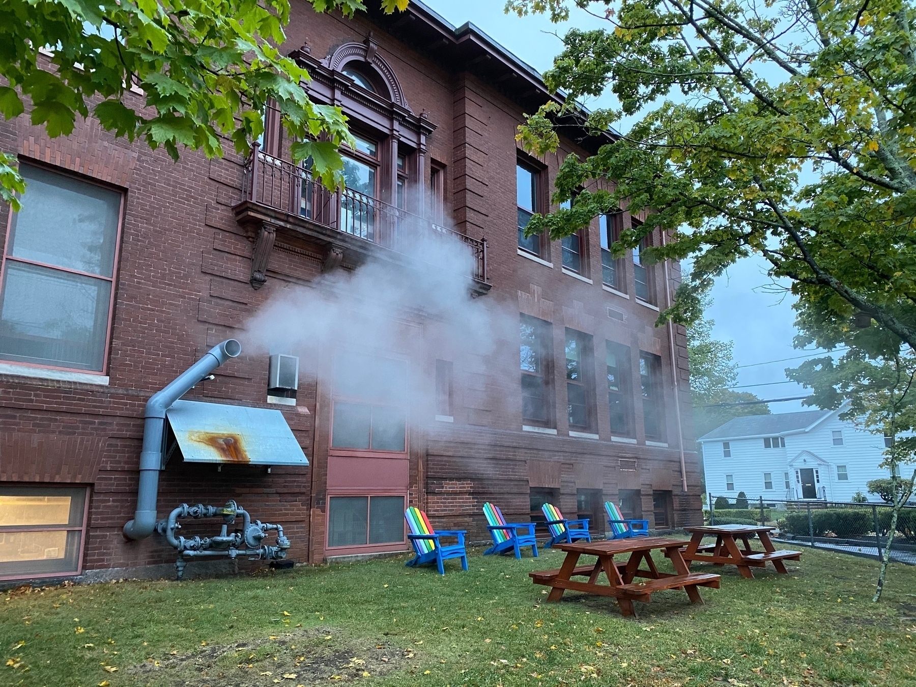 The side of a brick building with steam escaping from some pipes on the side, with picnic tables and adirondack chairs chairs on the lawn in front.