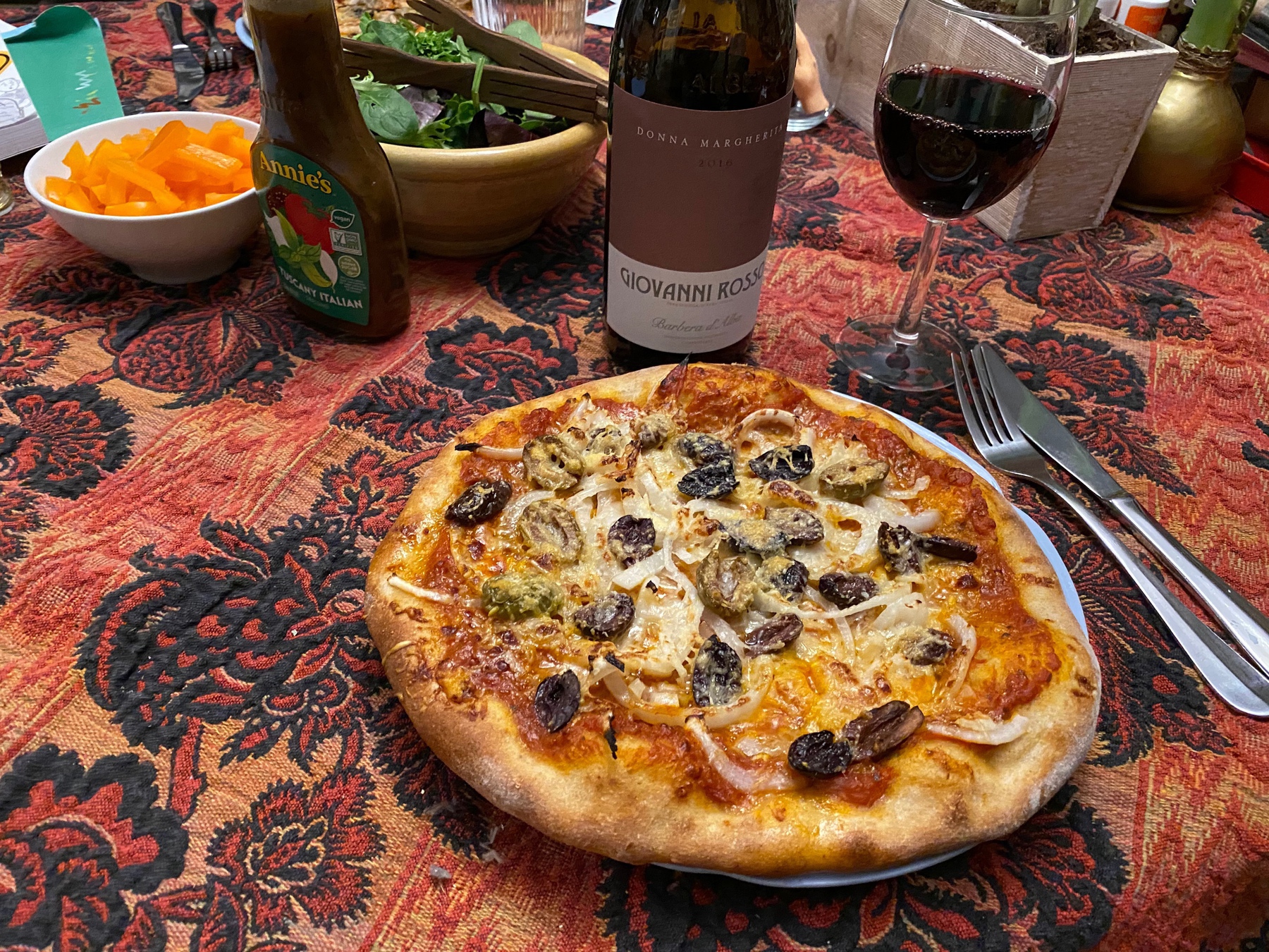 A small pizza on a plate with a glass of wine, a wine bottle, and a small salad in the background.